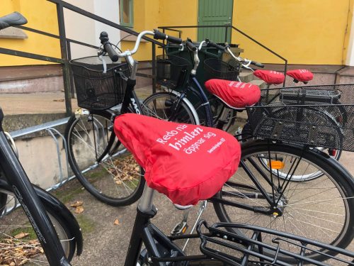 Ett antal cyklar i ett cykelställ, alla med röda cykelsadelskydd.